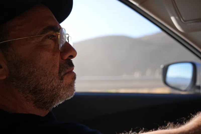 A middle-aged bearded man wearing a black cap and gold-framed glasses, in profile, driving or riding through an arid, mo