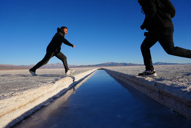 Two people leaping or running across opposite sides of a narrow water-filled channel cut into a salt flat, likely the Sa