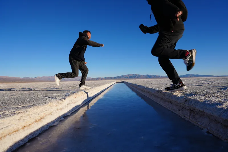 Two people leaping over or across a brine channel cut into a salt flat, likely the Salinas Grandes in northwest Argentin