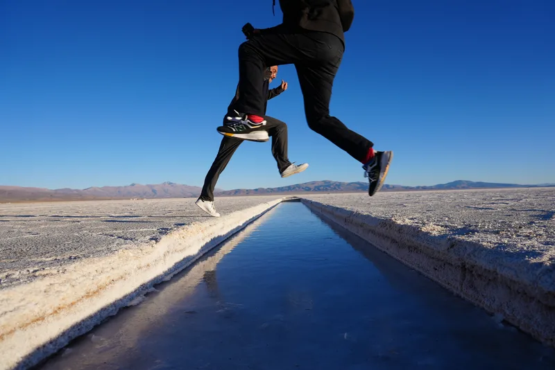 A person mid-leap over a narrow brine extraction channel on a South American salt flat, likely the Salinas Grandes in Ar