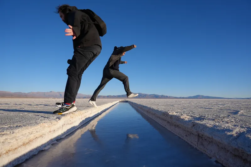Two figures leaping over or across a brine channel cut into the Salinas Grandes salt flat, likely northwestern Argentina