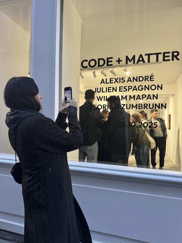 A woman photographs a gallery opening through a street-facing window, with the exhibition title 'CODE + MATTER' printed 