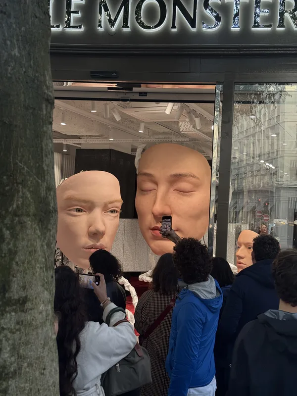 A crowd photographing oversized sculptural human heads in a Paris boutique window display, partially observed from behin