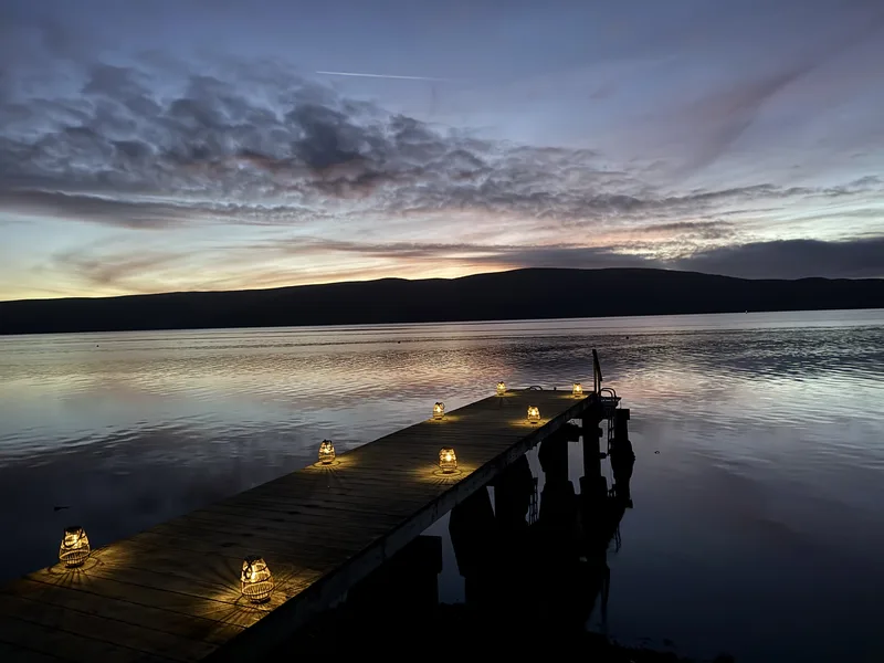 A wooden dock lined with lit lanterns extending over a calm lake at twilight, with a mountain silhouette and layered sky