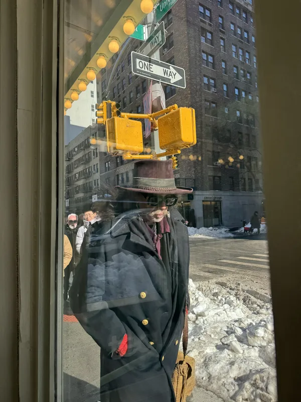 A figure in a theatrical, anachronistic outfit — wide-brimmed hat, long dark military-style coat with gold buttons, red 