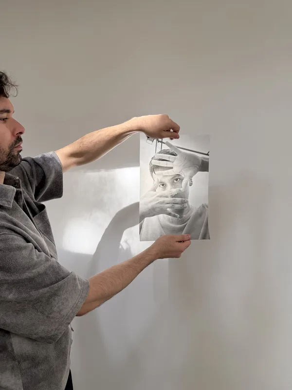 A bearded man in profile holding a black-and-white photographic print of a face obscured by hands