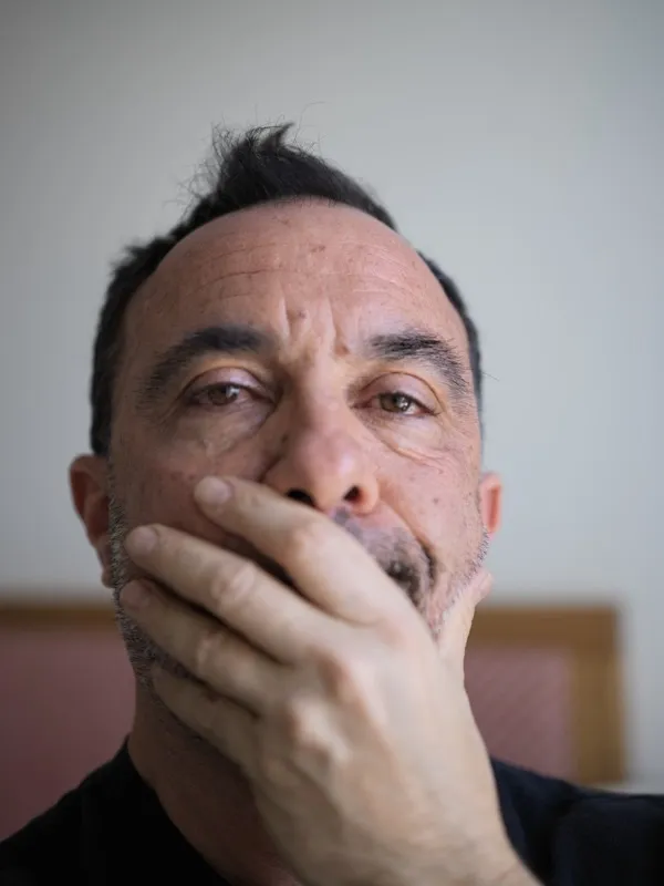 A middle-aged man of Mediterranean or Middle Eastern appearance, hand raised to cover his mouth, eyes directed upward to