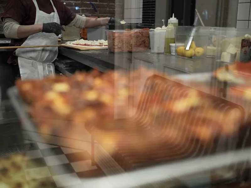 A pizza maker in white apron and black gloves preparing pies at a commercial pizzeria counter, viewed through a glass di