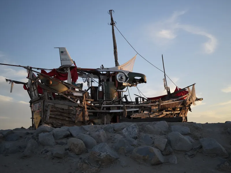 A large-scale assemblage constructed from salvaged materials — timber planking, rope rigging, red fabric, and the empenn