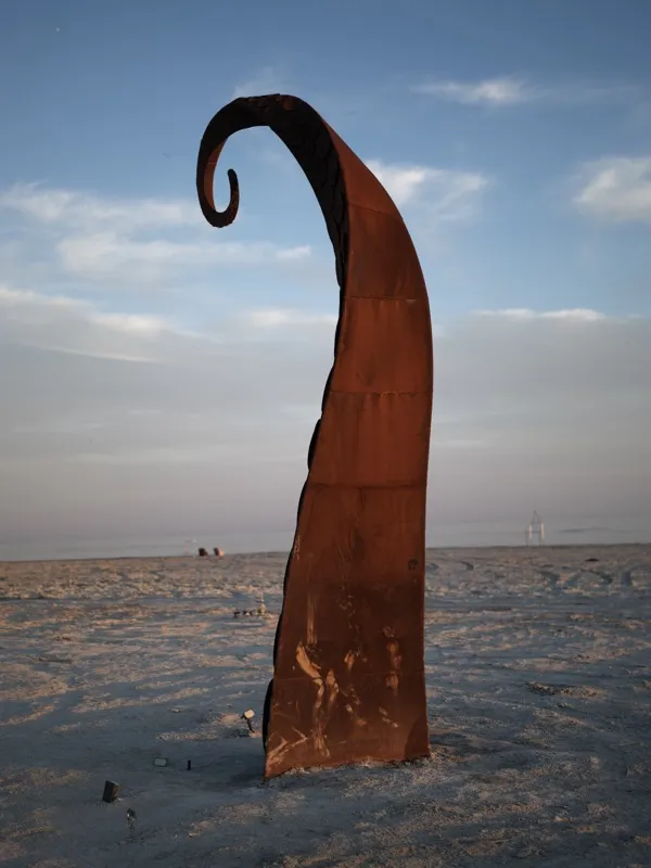 A Corten steel abstract sculpture, vertically oriented, terminating in a tight spiral curl, installed on a flat arid pla