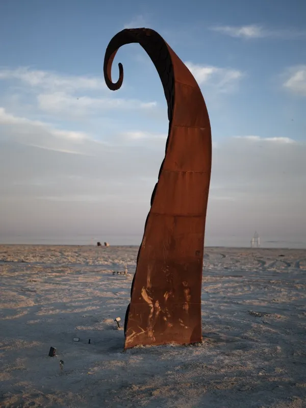 A large-scale Corten steel public sculpture, heavily oxidized, with a curling upper terminus, installed on a coastal sal