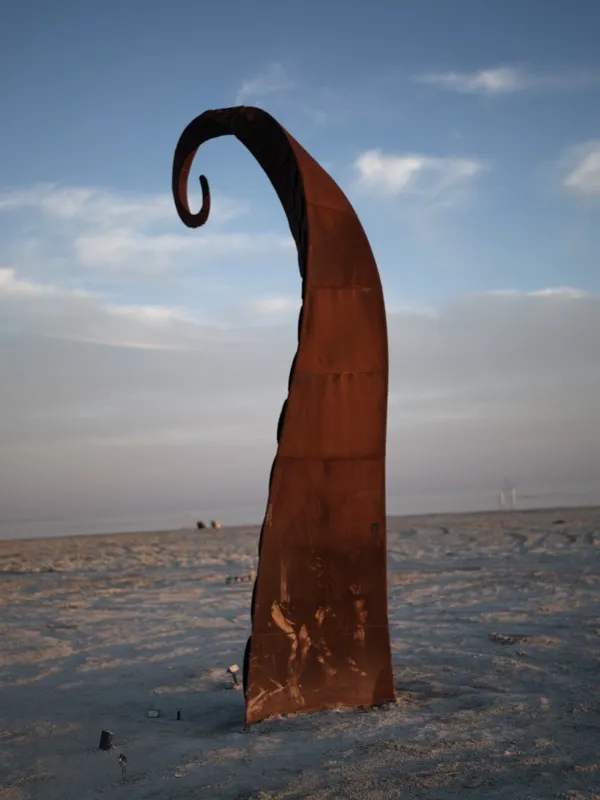 A Corten weathering steel sculpture, freestanding on a flat arid plain, photographed at golden hour