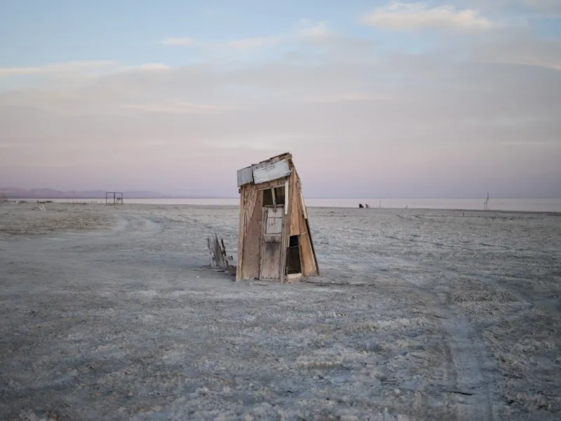 A partially collapsed wooden outhouse, tilting severely, standing alone on a bleached salt flat at dusk