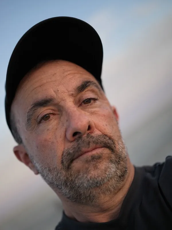 A middle-aged man, olive-complexioned, salt-and-pepper beard, black baseball cap, dark clothing — photographed in late a