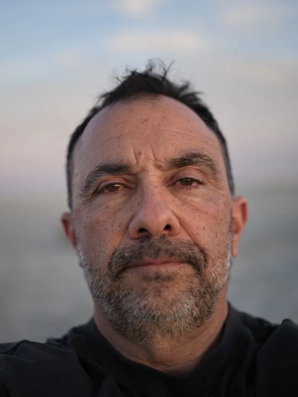 A middle-aged Mediterranean or Middle Eastern man, dark hair with gray at the temples, salt-and-pepper stubble, wearing 