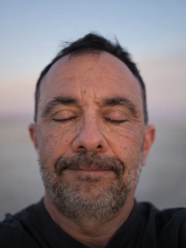 A middle-aged man, roughly late forties to mid-fifties, eyes closed, photographed at close range against an open-sky bac