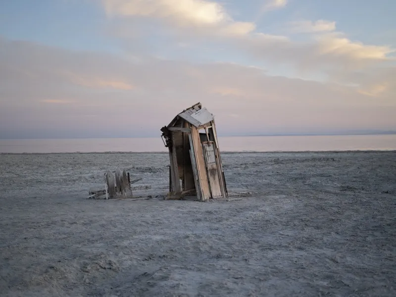 A collapsed wooden outhouse tilting on a dried salt flat at twilight