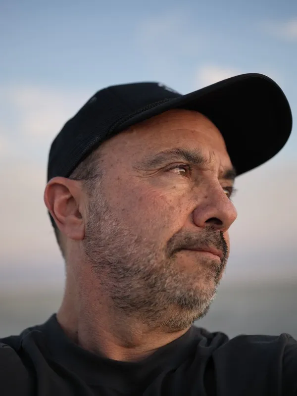 A middle-aged man in a black baseball cap and black hoodie, photographed in three-quarter profile at dusk