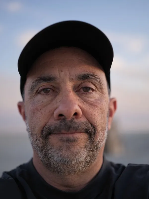 Middle-aged man, approximately 50-60 years old, dark complexion, salt-and-pepper stubble, wearing a black baseball cap a