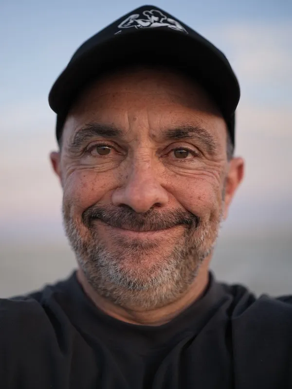 Middle-aged man, Mediterranean or Middle Eastern features, salt-and-pepper beard, black baseball cap with a logo, black 