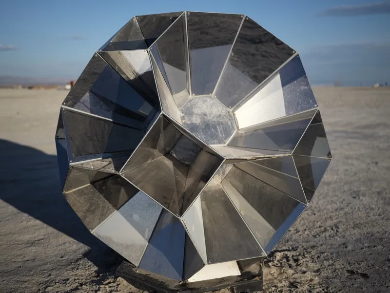 A large polyhedral stainless steel sculpture with spiral-arranged faceted panels, resting on the surface of a dry lakebe