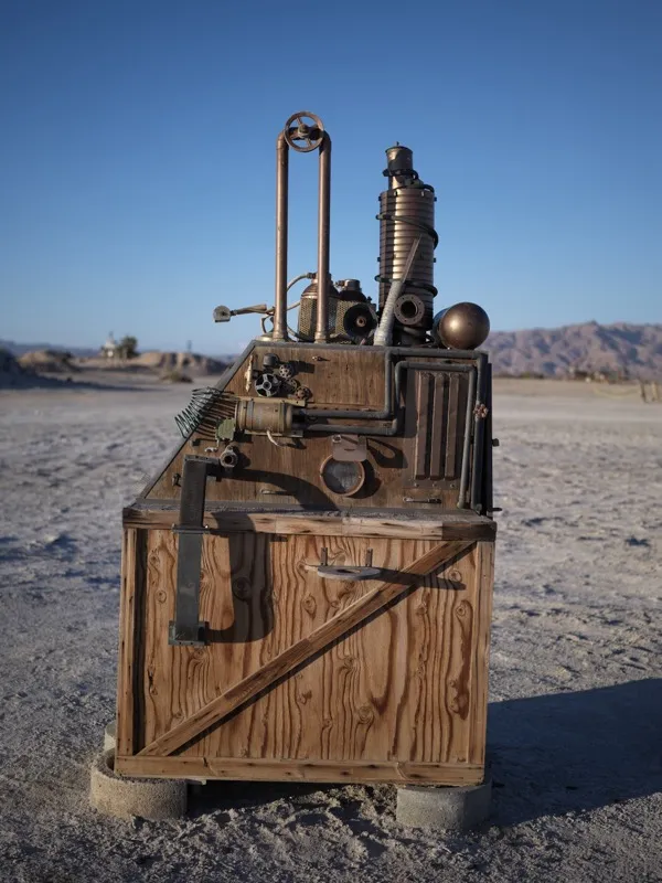 A fabricated sculptural machine — assemblage of weathered plywood crating, copper pipe, corrugated metal cylinders, salv