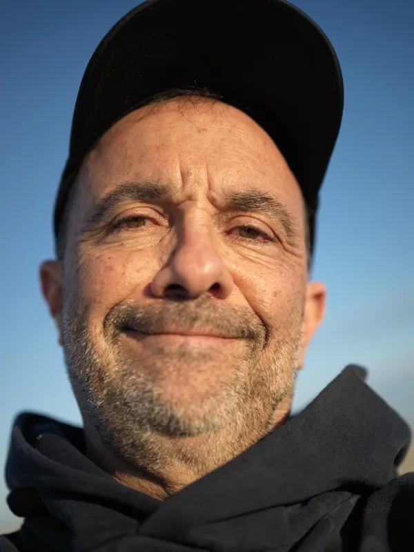 A middle-aged man, Mediterranean or Middle Eastern appearance, wearing a black baseball cap and black jacket, outdoors i