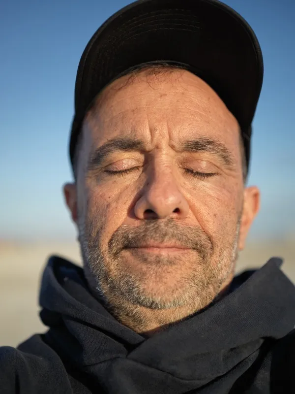 A middle-aged man, eyes closed, face turned slightly upward toward late-afternoon sunlight, wearing a black baseball cap
