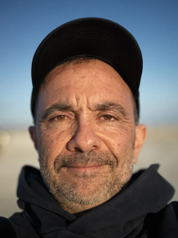 A middle-aged man, Mediterranean or Middle Eastern features, wearing a black baseball cap and dark jacket. Likely late f