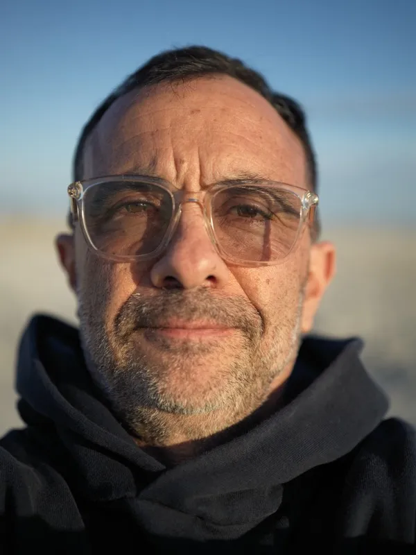A middle-aged man, mid-fifties, wearing clear-framed rectangular glasses and a dark navy hooded garment. Salt-and-pepper