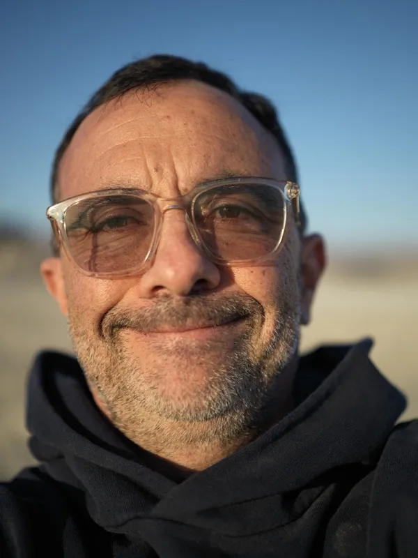 A middle-aged man, glasses, stubble, dark hoodie, photographed from close range outdoors at golden hour.