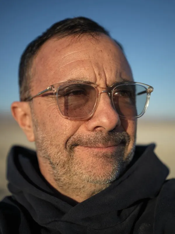 A middle-aged man, salt-and-pepper beard, translucent-framed glasses, dark hoodie, photographed at close range in an ari