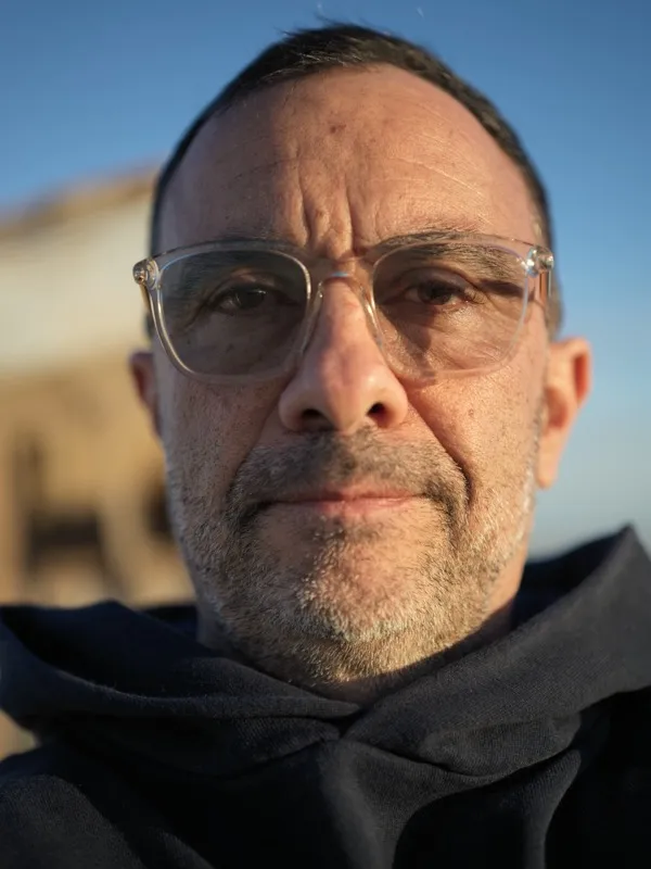 A middle-aged man, dark clothing, clear-framed glasses, salt-and-pepper stubble, photographed at close range outdoors at
