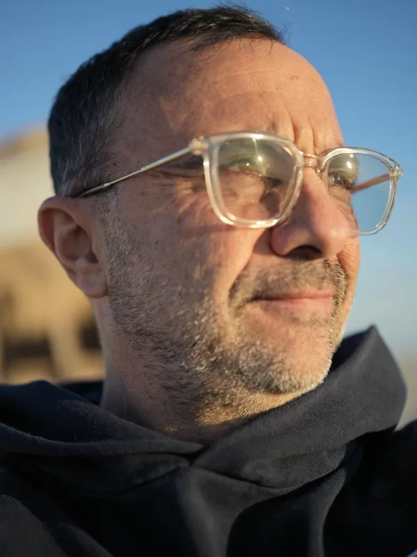A middle-aged man, balding, with salt-and-pepper stubble and clear-framed rectangular glasses, photographed outdoors in 