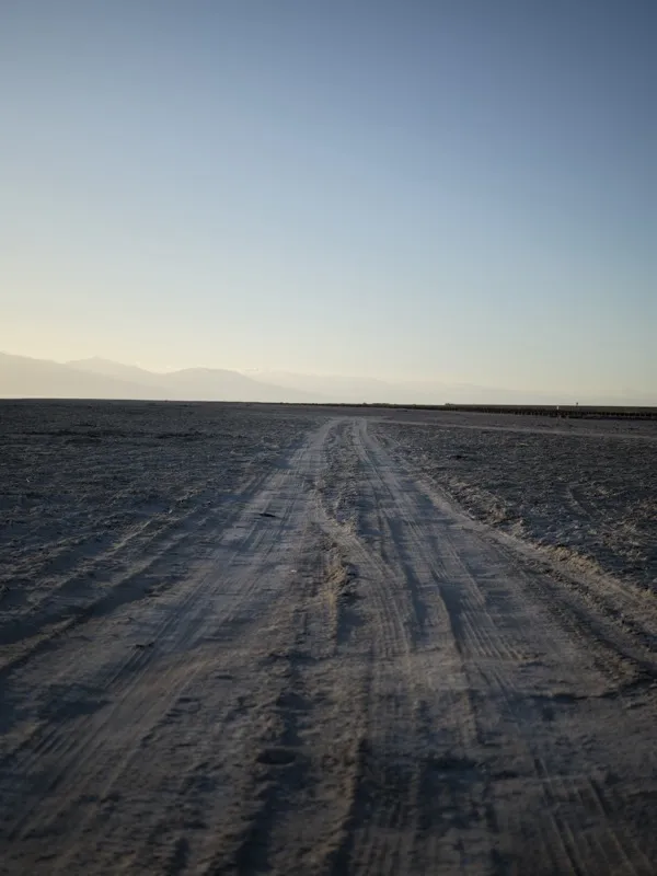 An unpaved desert road or dry lakebed track, marked by vehicle tire impressions, extending to the horizon across a flat,