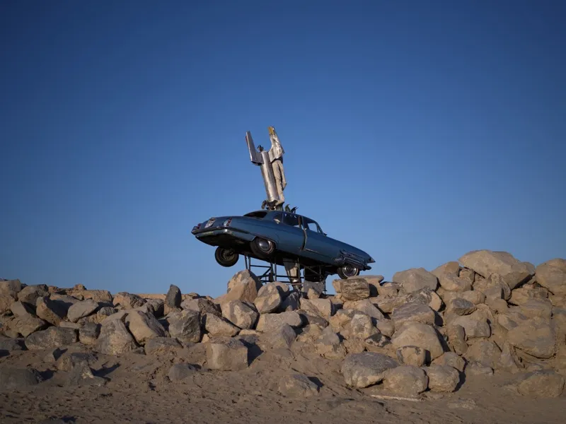 A vintage E-Type Jaguar or similar 1960s coupe, elevated on metal scaffold above a desert boulder field, with a rocket o