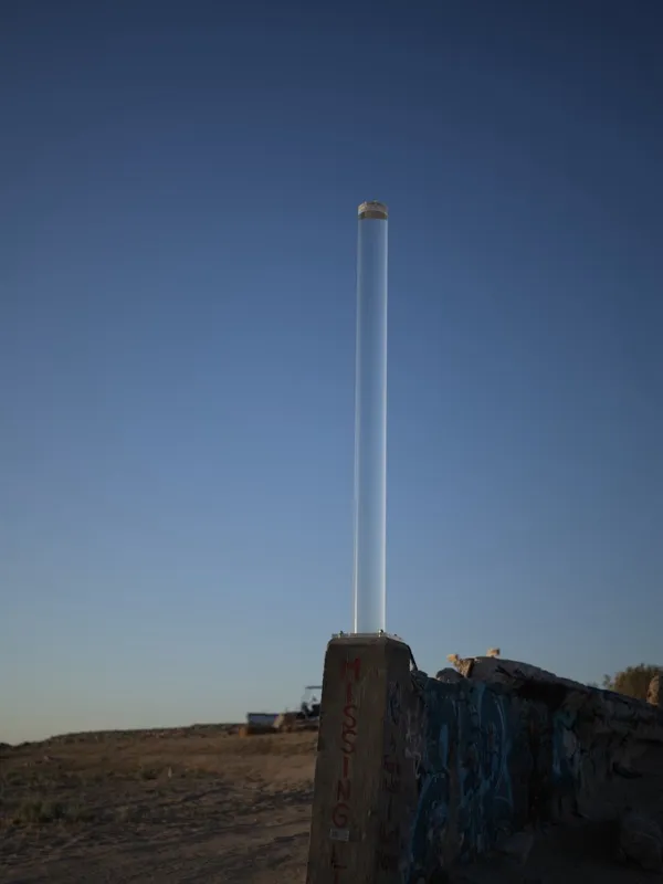 A fluorescent tube mounted vertically on a deteriorating graffiti-marked concrete wall in an arid, semi-abandoned landsc