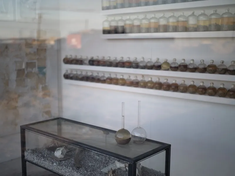 A contemporary art installation involving laboratory glassware and geological or sedimentary materials, photographed thr