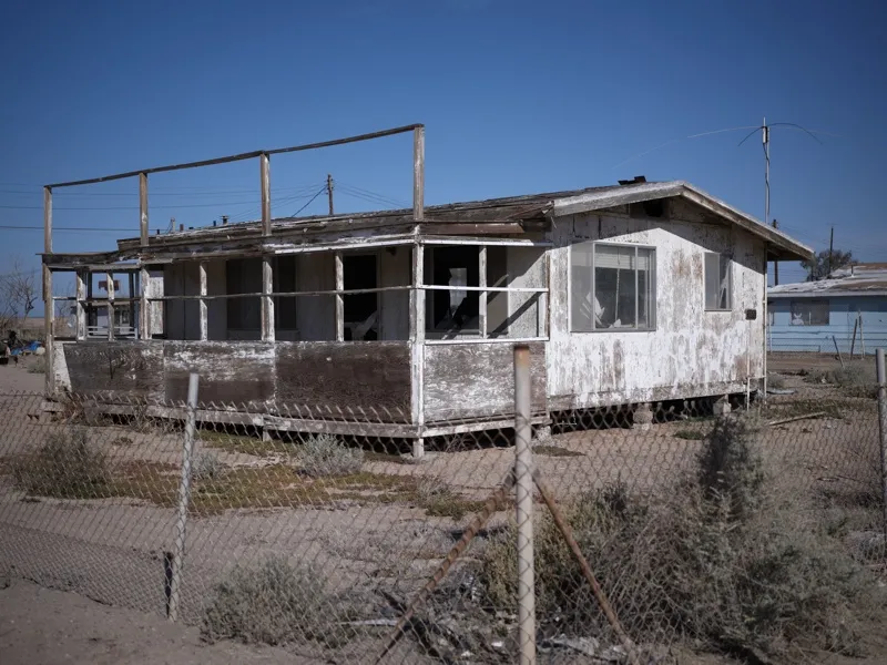 An abandoned single-story wood-frame dwelling in a desert community, surrounded by chain-link fencing and sparse scrub v