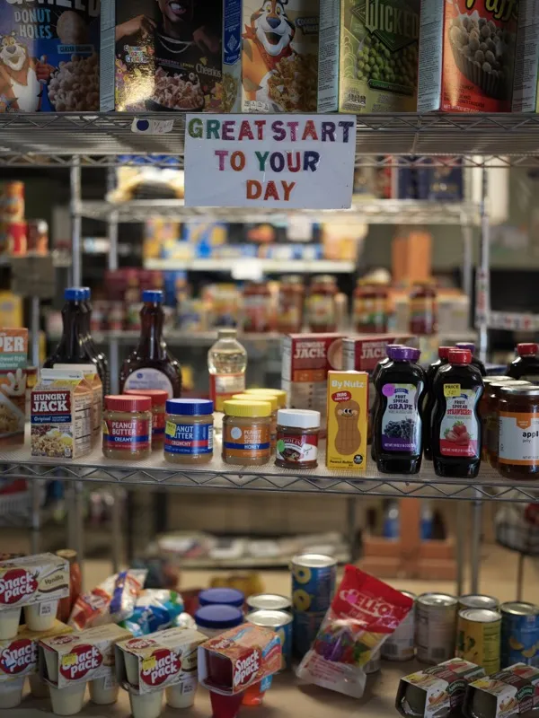 A food bank or community pantry shelf stocked with breakfast staples — peanut butter, jelly, syrup, cereals — topped by 