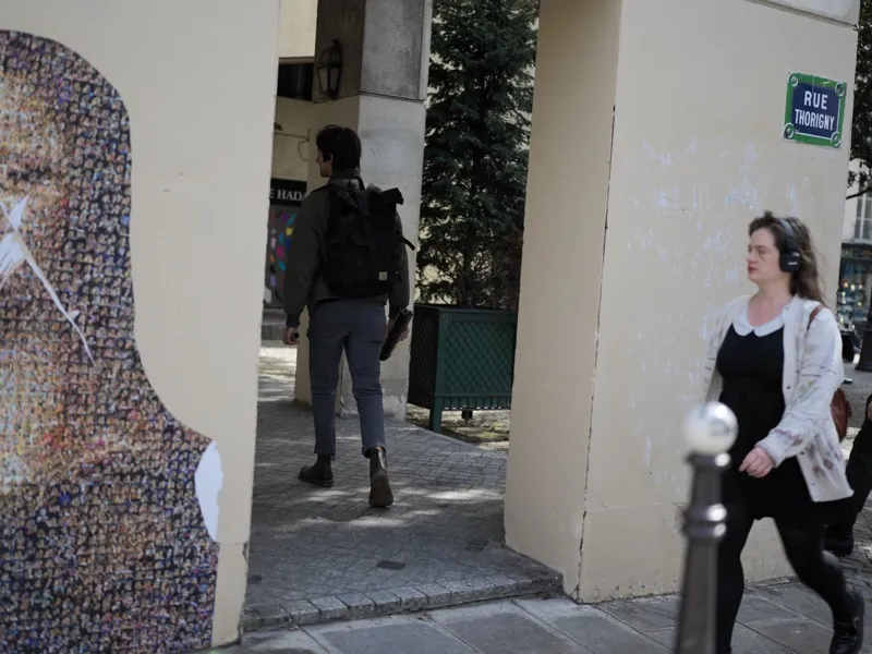Two Parisian pedestrians in opposing motion, framed between a monumental mosaic portrait and a stone archway on Rue Thor