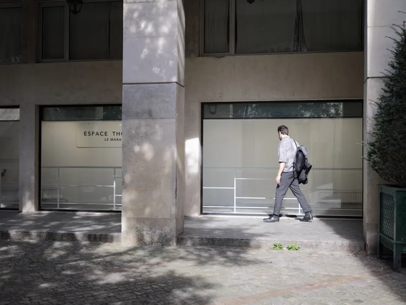 A man in business-casual dress walking past the facade of a cultural space — likely a gallery or théâtre — in Paris's Ma