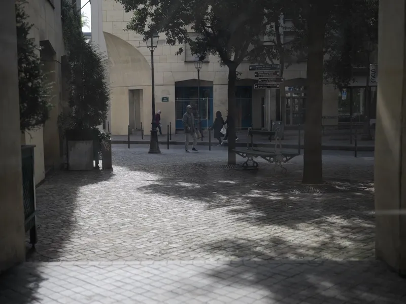 A French urban plaza viewed through a sheltered passageway, populated by scattered pedestrians in morning light