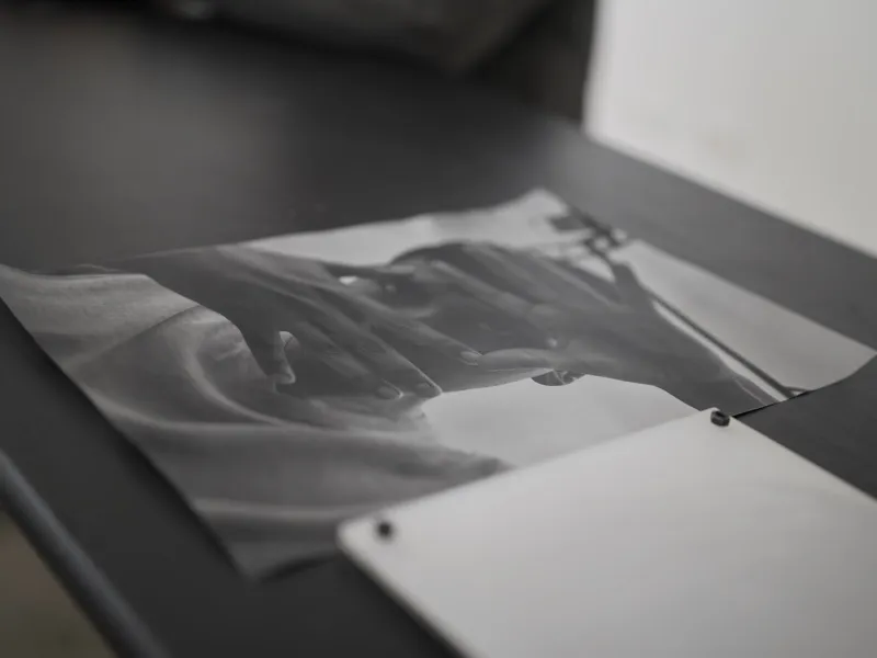 A monochrome photographic print depicting two hands in intimate contact, lying on a dark work surface beside a blank she