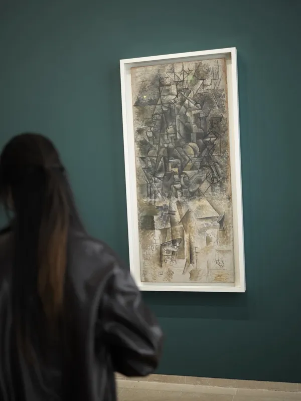 A young woman with dark hair in a leather jacket stands before a small Analytic Cubist painting in a museum gallery.