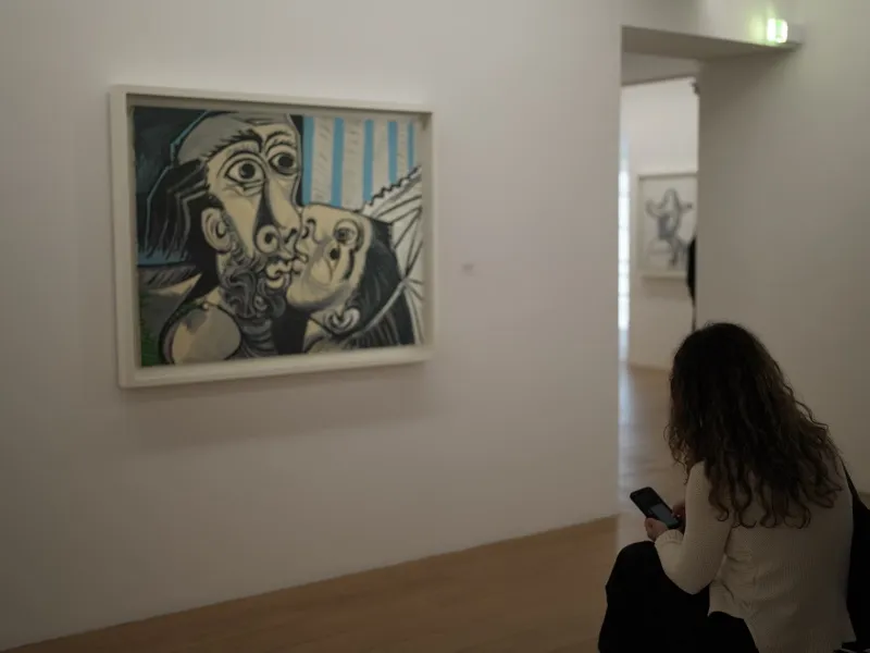 A young woman seen from behind, seated in a gallery, looking at her phone while a large Cubist painting of an embrace ha