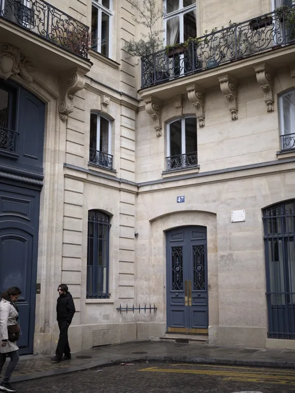 The corner facade of a Haussmann-era Parisian apartment building, number 47, with two pedestrians passing below