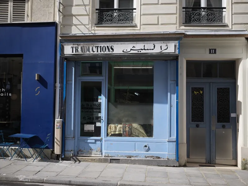 A small Parisian translation bureau, its storefront painted powder blue, bearing a bilingual sign in French and Arabic s