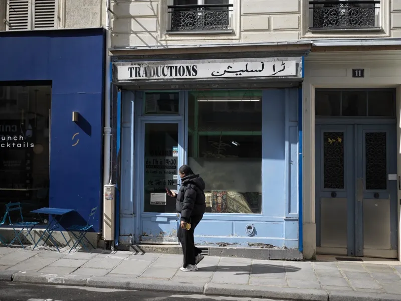 A solitary figure in all-black clothing, absorbed in a phone, standing before a bilingual French-Arabic translation shop
