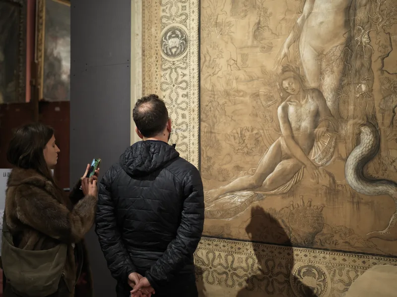 Two museum visitors — a woman photographing on her phone, a man standing with hands clasped behind his back — standing b